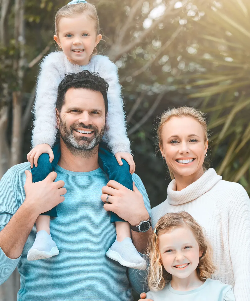 A smiling family outdoors, a young girl riding on her father's shoulders